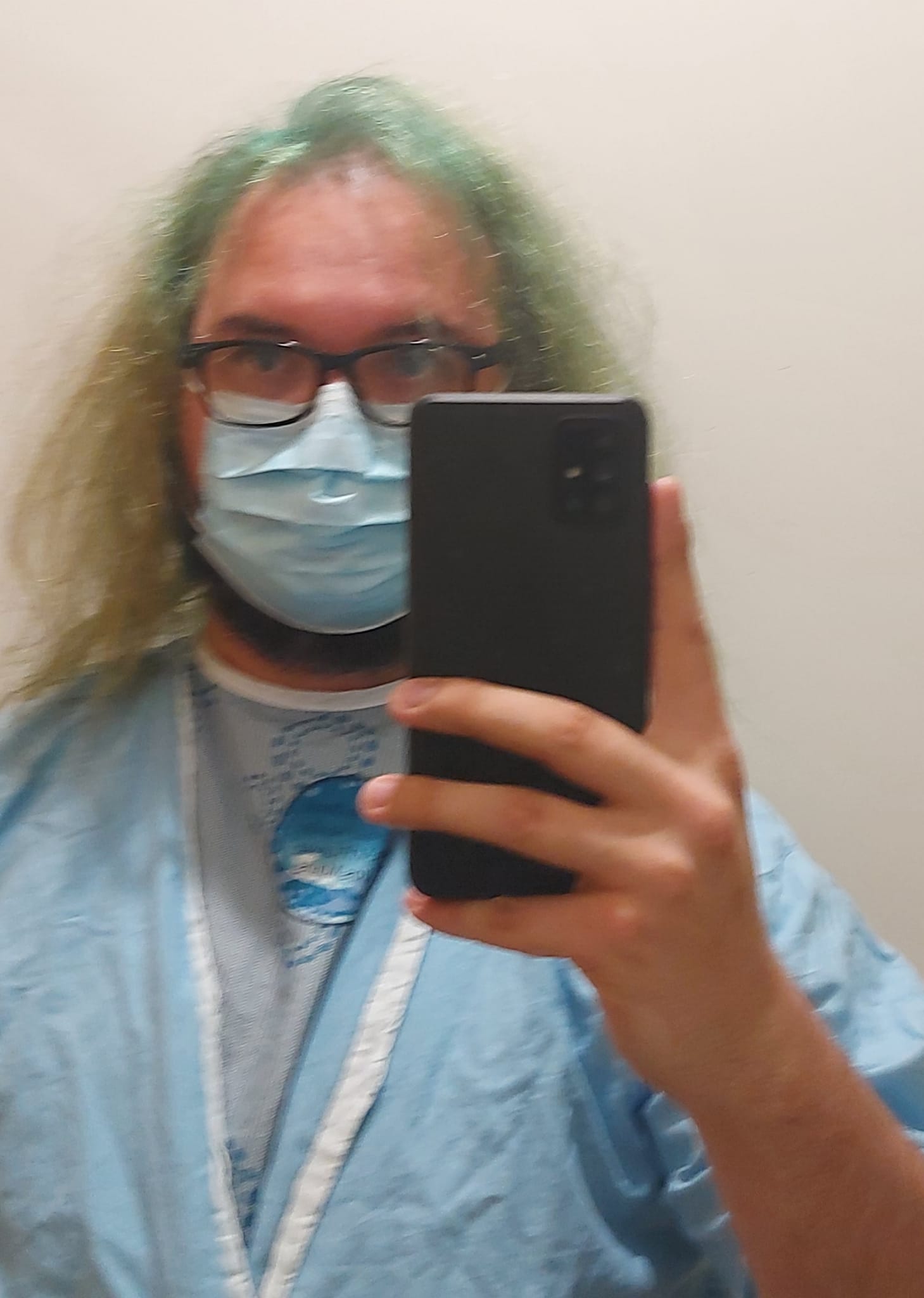 A man with green hair and a beard is wearing a hospital gown, phone visible in classic mirror selfie pose